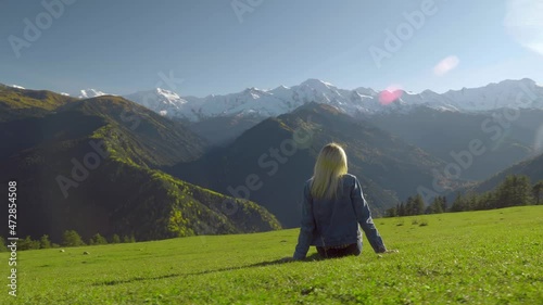 after resting on the top of the mountain after hiking, a female traveler sits down on the grass and admires nature, the view from behind, exploring natural attractions. georgia, svaneti kheshkili. photo