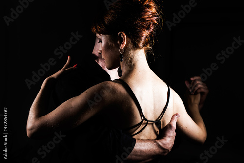 Young couple - a man and a woman wearing black dancing in the dark; the lady stands with her back to the camera.