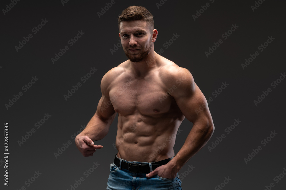 Waist up portrait of bearded shirtless bodybuilder looking at the camera while posing over the black wall. Stock photo Waist up portrait of bearded shirtless bodybuilder looking at the camera while posing over the black wall. Stock photo