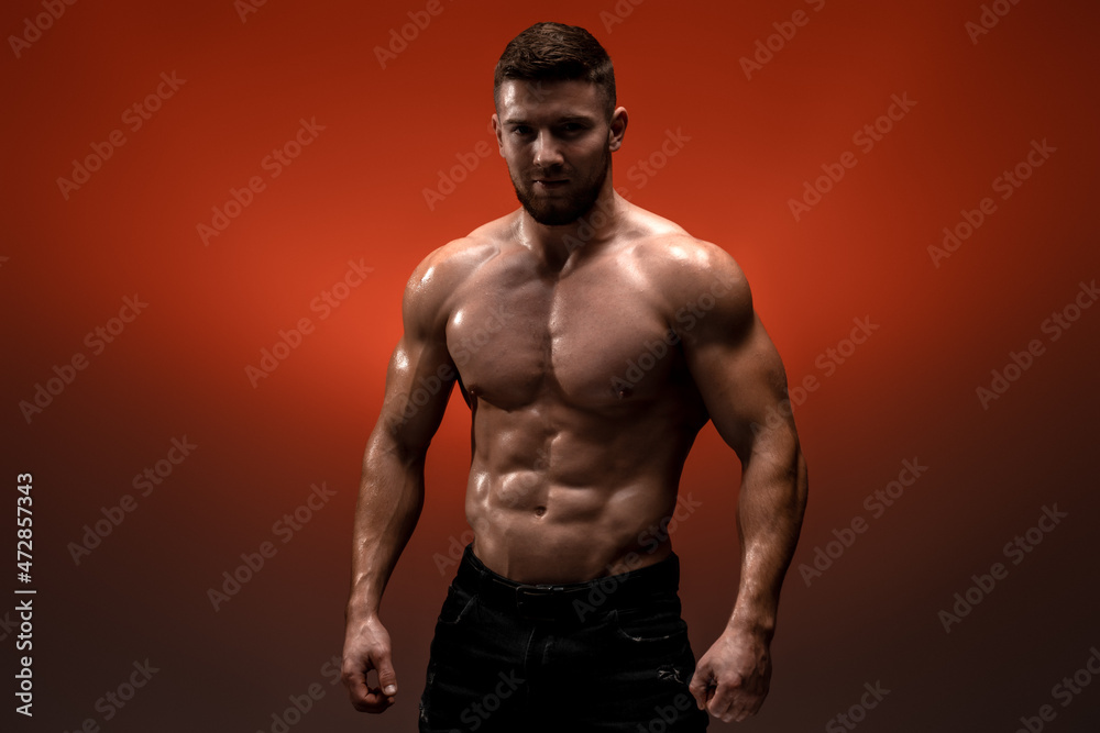 Fototapeta premium Serious sportsman is looking at the camera while standing at the studio. His torso is naked. Isolated on red background
