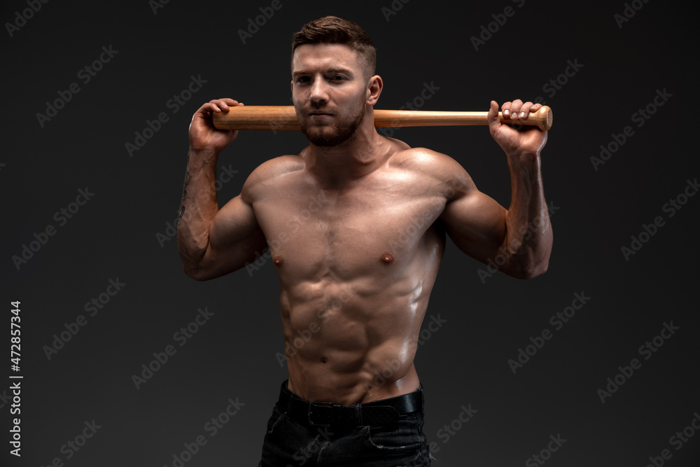 Strong and fit man bodybuilder with baseball bat shows abdominal muscles. Sporty muscular guy athlete. Sport and fitness concept Strong and fit man bodybuilder with baseball bat shows abdominal muscles. Sporty muscular guy athlete. Sport and fitness concept