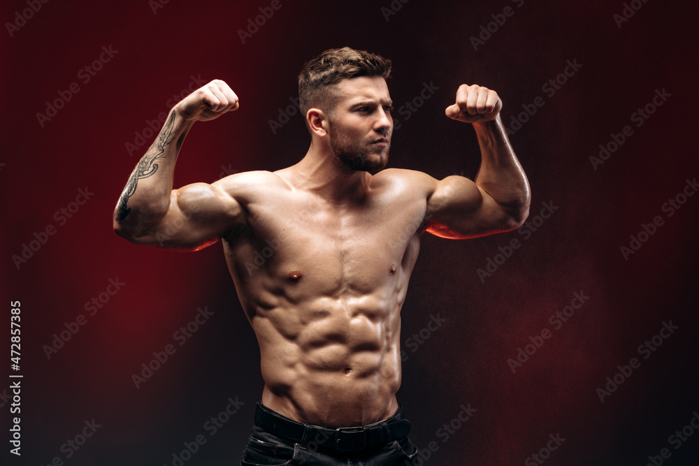 Obraz premium Young athletic man with a naked torso showing his biceps isolated on a red background. Bodybuilding and strong people concept