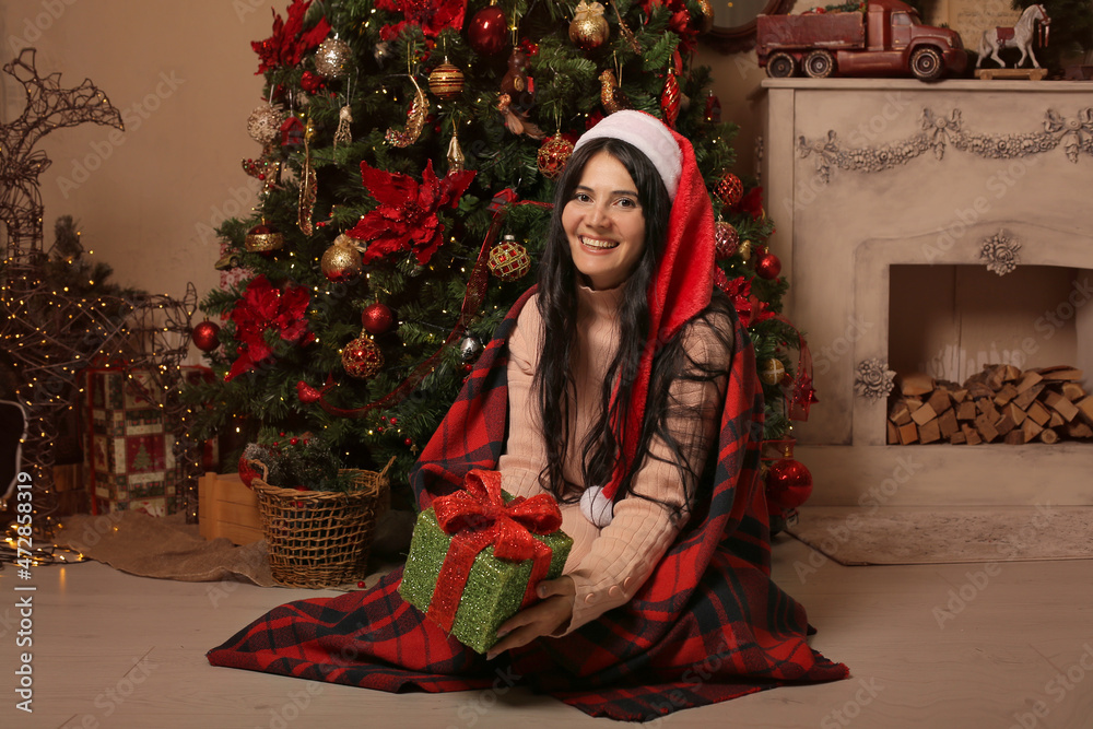 a brunette girl in a Santa Claus hat in pink pajamas is sitting covered with a plaid plaid by the fireplace in her hands a gift box Christmas tree new year