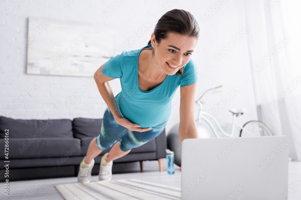 happy pregnant woman with sportswear working out near laptop in living room.