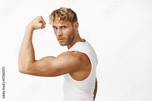 Portrait of handsome strong male athlete, fitness instructor flexing biceps, showing biceps on arm and looking confident at camera, white background
