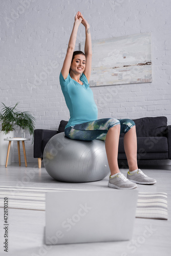 positive pregnant woman with raised hands exercising on fitness ball and looking at laptop in living room.