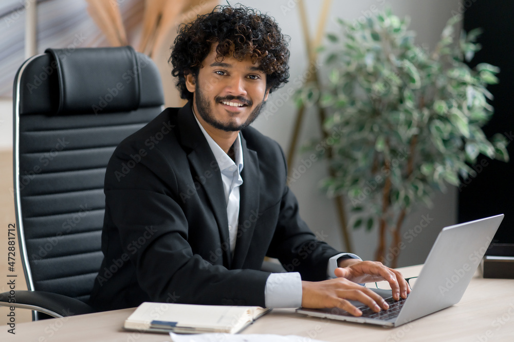 Portrait of a positive charismatic curly-haired young adult Indian businessman, wearing business attire, sitting at his desk in the office, looking at the camera with a friendly smile Portrait of a positive charismatic curly-haired young adult Indian businessman, wearing business attire, sitting at his desk in the office, looking at the camera with a friendly smile