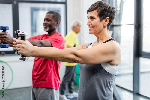 Elderly sportswoman training with dumbbells near african american friend in gym.