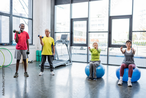 Multiethnic sportsmen training with dumbbells near sportswomen on fitness balls in gym.