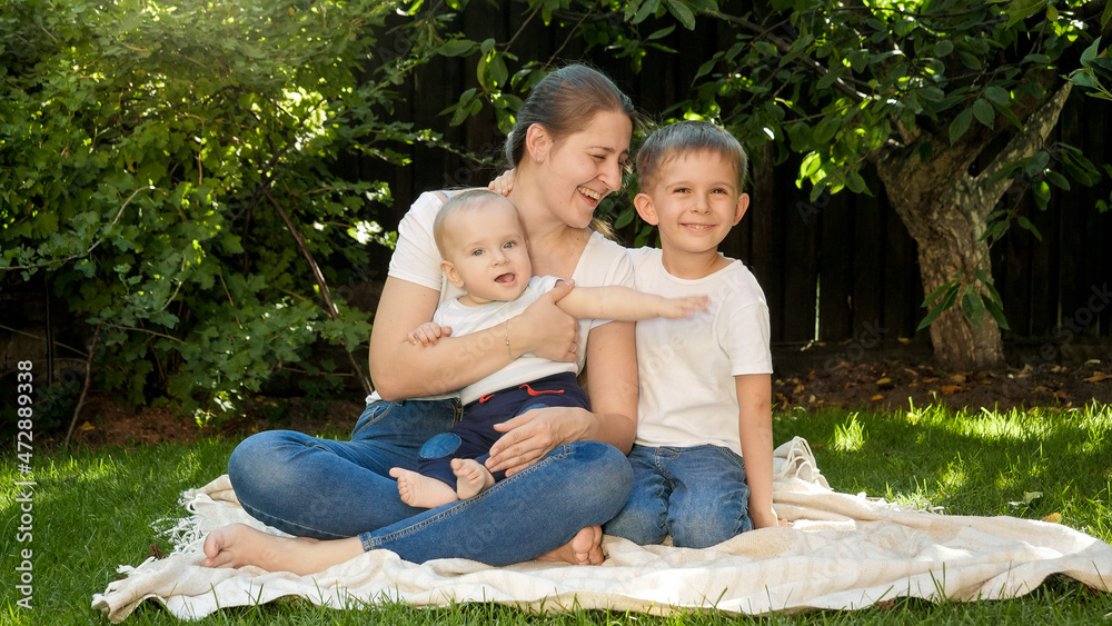 Older boy hugging his baby brother and mother sitting on grass in garden. Parenting, family, children development, and fun outdoors in nature. Older boy hugging his baby brother and mother sitting on grass in garden. Parenting, family, children development, and fun outdoors in nature.
