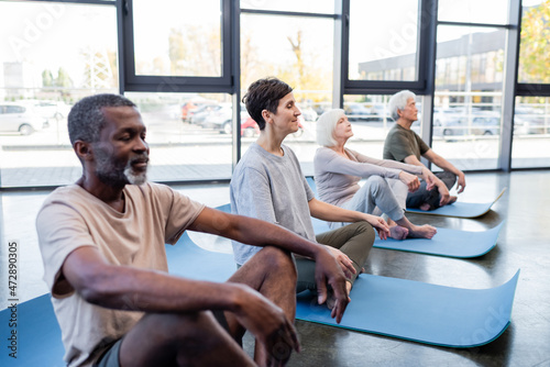 Senior woman meditating on yoga mat near interracial people in gym.