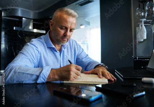 Charming middle aged entrepreneur, businessman writing in a notebook, planning projects, and making notes in a diary, sitting in front of a laptop monitor at home with styish interior.