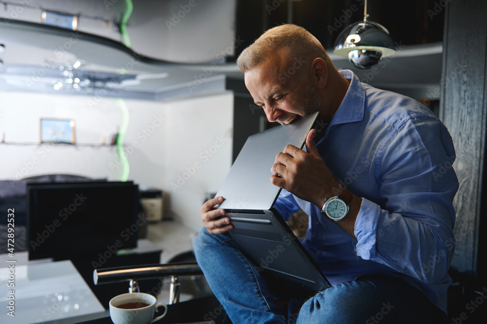 Middle aged angry Caucasian man, entrepreneur in casual clothes sitting on a table with a cup of coffee and holding a laptop in his hands, biting it with rage