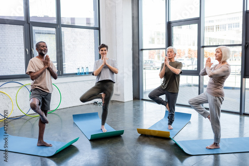 Interracial senior people standing in tree pose on yoga mats in sports center.