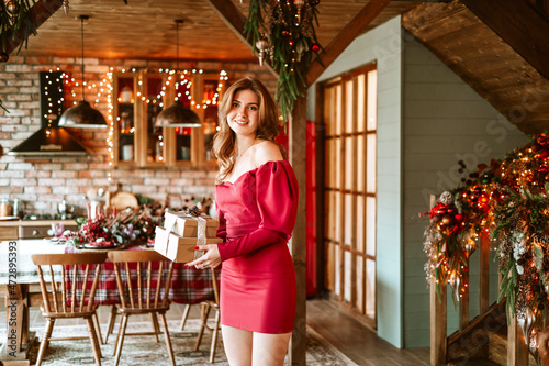 beautiful young woman in red dress stands near Christmas tree in living room decorated for celebration of Christmas and New Year and opens gift box with wrapped Christmas presents