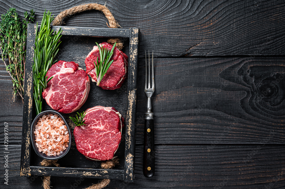 Fillet Mignon tenderloin raw meat veak steaks in wooden tray with herbs. Black wooden background. Top view. Copy space