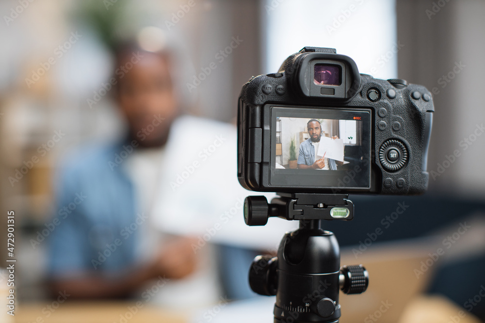 African american male freelancer with documents in hands talking during online concefere on blur background. Focus on modern digital camera.