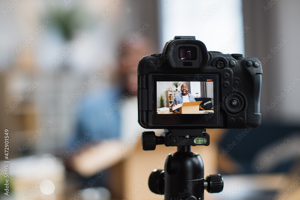 African american man sitting at desk and opening box with ordered products. Male influencer filming unpacking process on video camera. Concept of purchases, blogging and delivery.