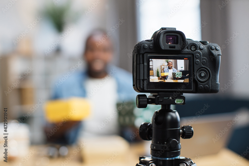 Blur background of african american blogger filming video while unpacking gift boxes at home. Focus on screen of modern camera. Freelance and technology concept.