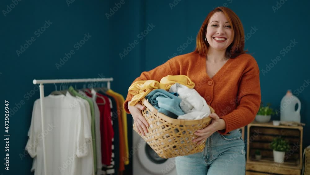 Young redhead woman smiling confident holding basket with clothes at laundry room