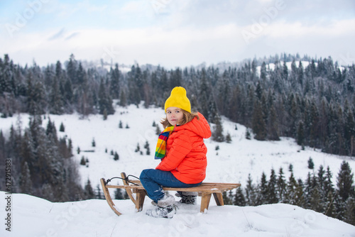 Portrait of happy little kid wearing knitted hat, scarf and sweater. Kid boy enjoying a sleigh ride. Child sledding riding a sleigh outdoors in snow. Winter knitted kids clothes.