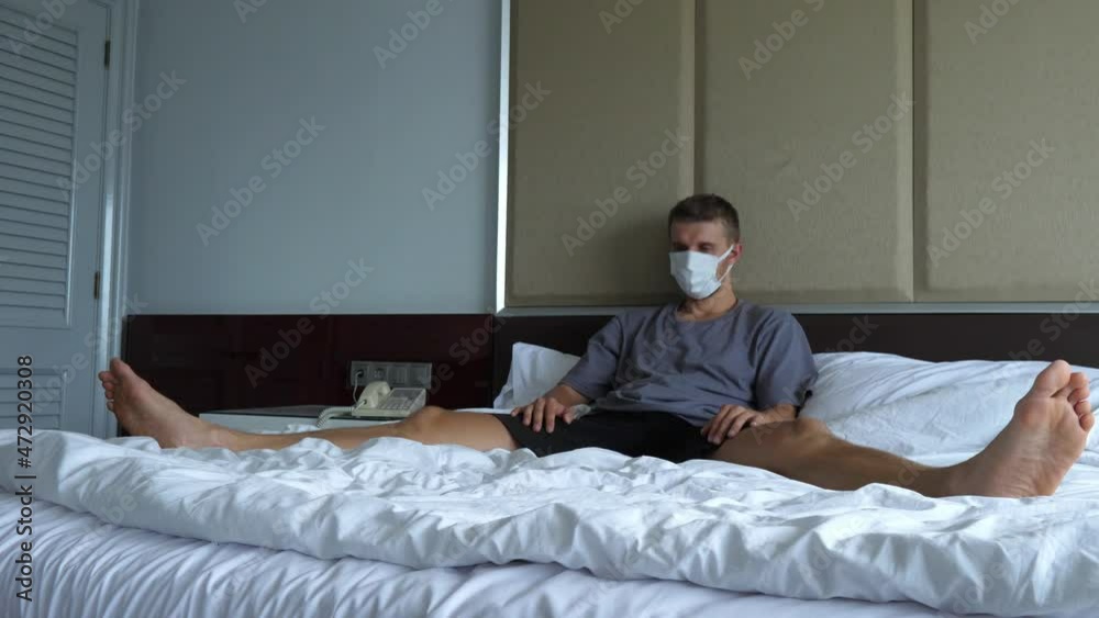 Bored man in a protective face mask quarantining, sitting on the bed and banging his head, staying at home during a new pandemic wave Bored man in a protective face mask quarantining, sitting on the bed and banging his head, staying at home during a new pandemic wave