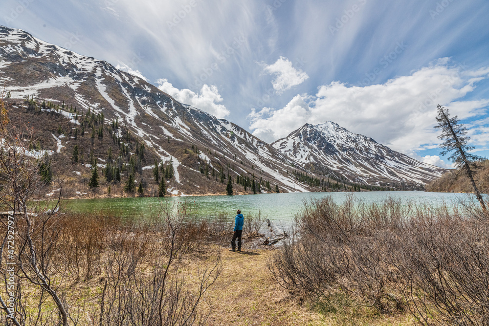 Hiker in blue jacket standing beside a isolated, wilderness lake in northern Canada during spring time, May. 