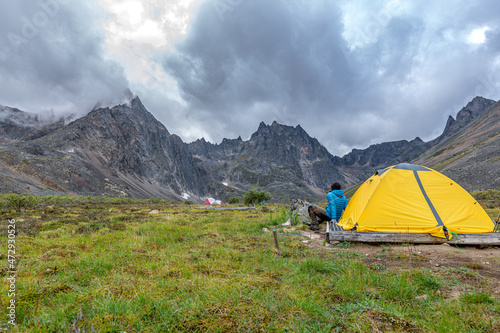 Camping area of Grizzly Lake in northern Canada during summer time, August with man hiker sitting beside yellow tent and epic mountains in background.