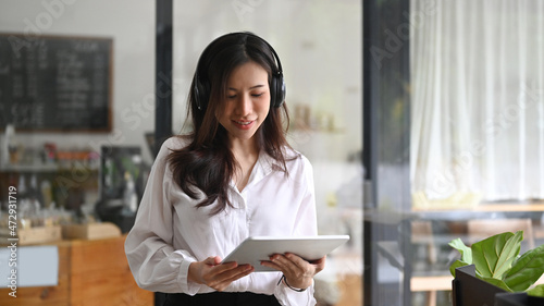 Positive asian woman wearing headphone and using digital tablet in coffee shop.