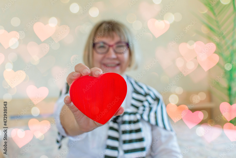Senior woman Valentine's Day with a heart in her hands on the background of her bedroom at home. love and old people concept. Senior Citizen's Day. Senior woman Valentine's Day with a heart in her hands on the background of her bedroom at home. love and old people concept. Senior Citizen's Day.