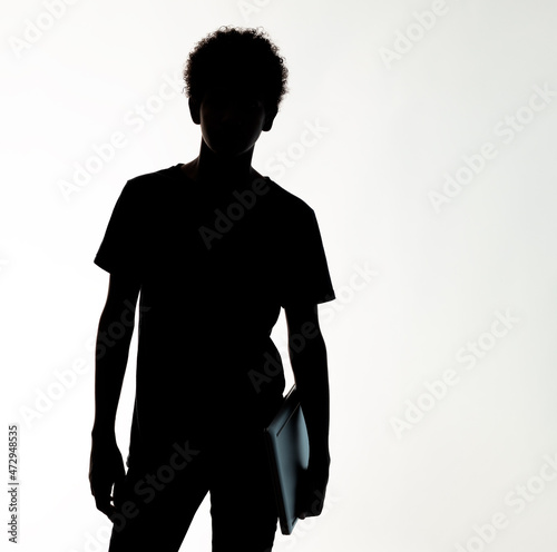silhouette a teenage boy with afro hair holding a laptop with his left hand, against the light
