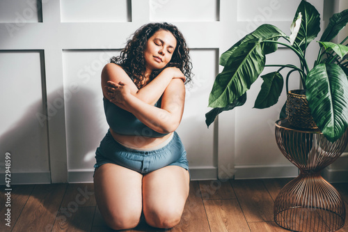 Curly haired overweight young woman in blue top and shorts with satisfaction on face accepts curvy body shape in stylish bedroom Fototapeta