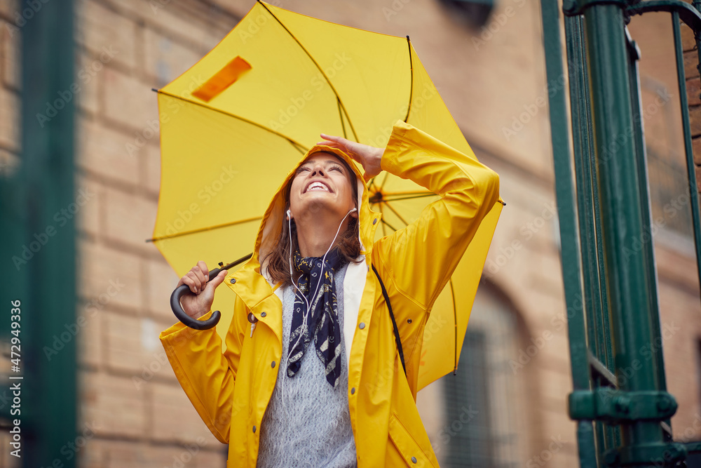 A young cheerful woman with a yellow raincoat and umbrella who is enjoying while listening to the music and walking the city on a rainy day. Walk, rain, city - obrazy, fototapety, plakaty 
