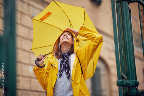 Fototapeta A young cheerful woman with a yellow raincoat and umbrella who is enjoying while listening to the music and walking the city on a rainy day