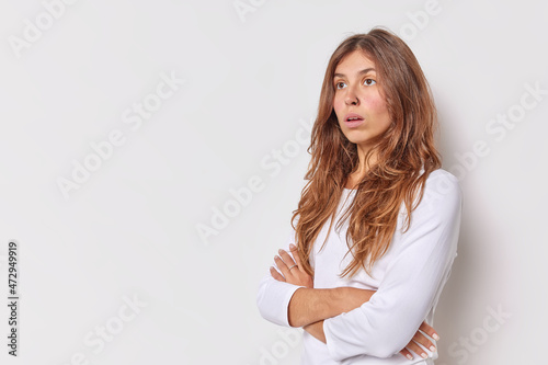 Photo of thoughtful long haired woman has surprised expression keeps arms folded concentrated somewhere with worried look poses against white backgrond with blank space for your promotion or text