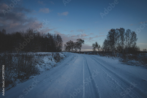 rarely used country road goes forward to forest, snow covered ground, road is slippery and dangerous, blue sky, winter sunset in Latvia, tire prints of only one vehicle