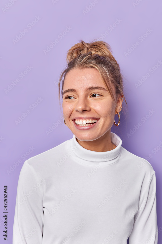Vertical shot of good looking young woman concentrated somewhere with merry expression notices something pleasant expresses sincere emotions wears casual white jumper isolated over purple wall Vertical shot of good looking young woman concentrated somewhere with merry expression notices something pleasant expresses sincere emotions wears casual white jumper isolated over purple wall