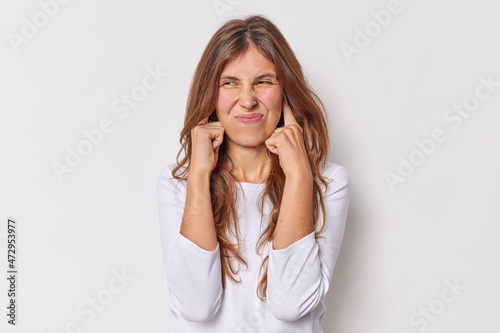 Young woman being disturbed by loud noise plugs ears with fingers frowns face avoids annoying sound which gives her headache dressed in casual jumper isolated over white background grimaces displeased