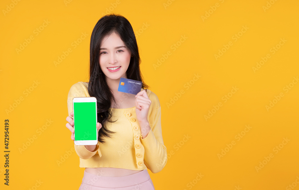 Cheerful smile asian woman holding smart phone and mockup credit card for internet banking while standing over isolated yellow background. Financial payment online shopping and money transfer concept.