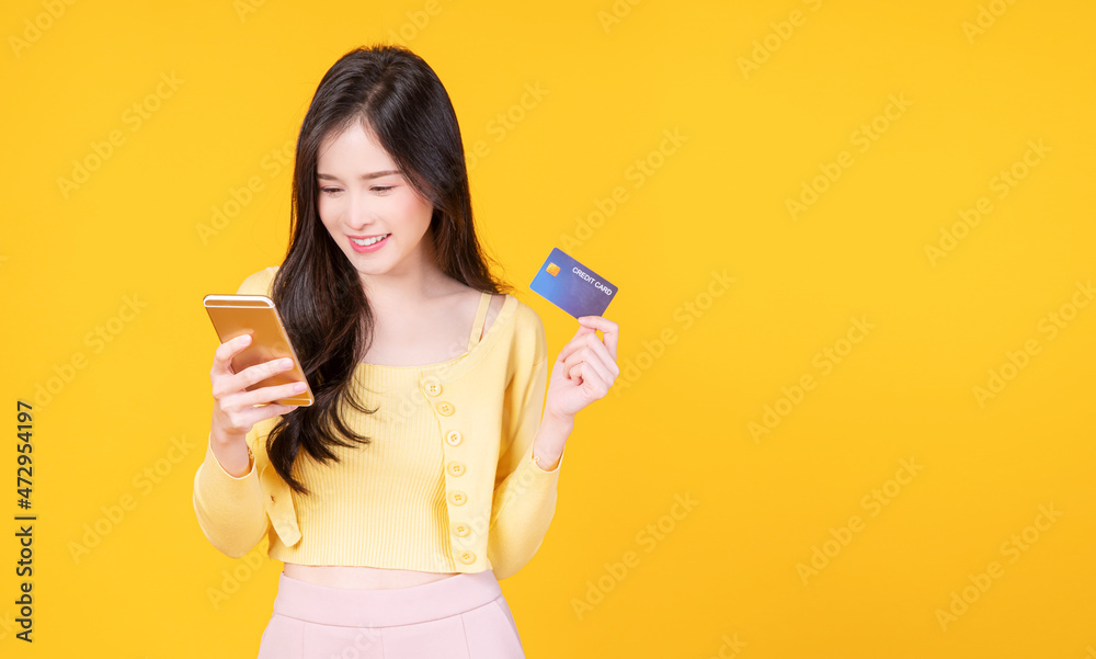 Cheerful smile asian woman holding smart phone and mockup credit card for internet banking while standing over isolated yellow background. Financial payment online shopping and money transfer concept. Cheerful smile asian woman holding smart phone and mockup credit card for internet banking while standing over isolated yellow background. Financial payment online shopping and money transfer concept.