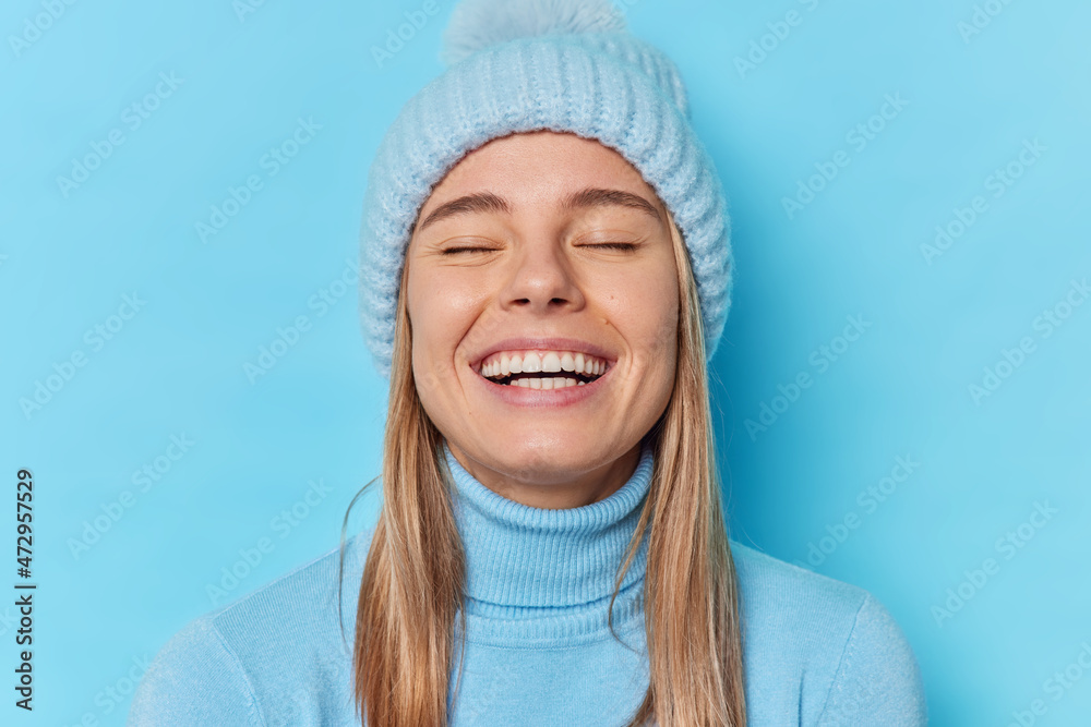 Portrait of happy sincere woman keeps eyes closed smiles broadly enjoys life to fullest laughs out loudly has fun dressed in casual poloneck and hat isolated over blue background. Emotions concept Portrait of happy sincere woman keeps eyes closed smiles broadly enjoys life to fullest laughs out loudly has fun dressed in casual poloneck and hat isolated over blue background. Emotions concept