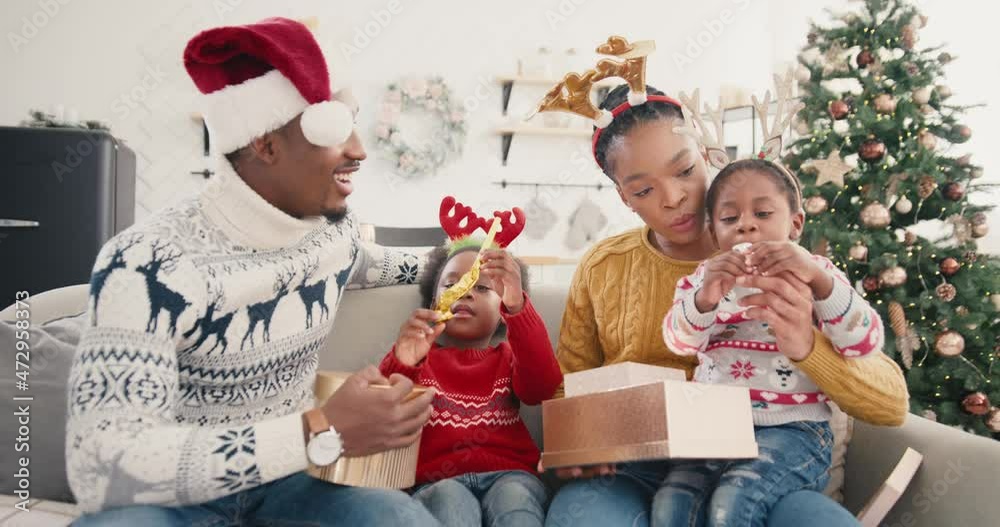 Portrait of curious African American kids open gift boxes while sitting with happy parents together at decorated home near pretty Christmas tree New Year Eve. Holidays celebration xmas present concept Portrait of curious African American kids open gift boxes while sitting with happy parents together at decorated home near pretty Christmas tree New Year Eve. Holidays celebration xmas present concept