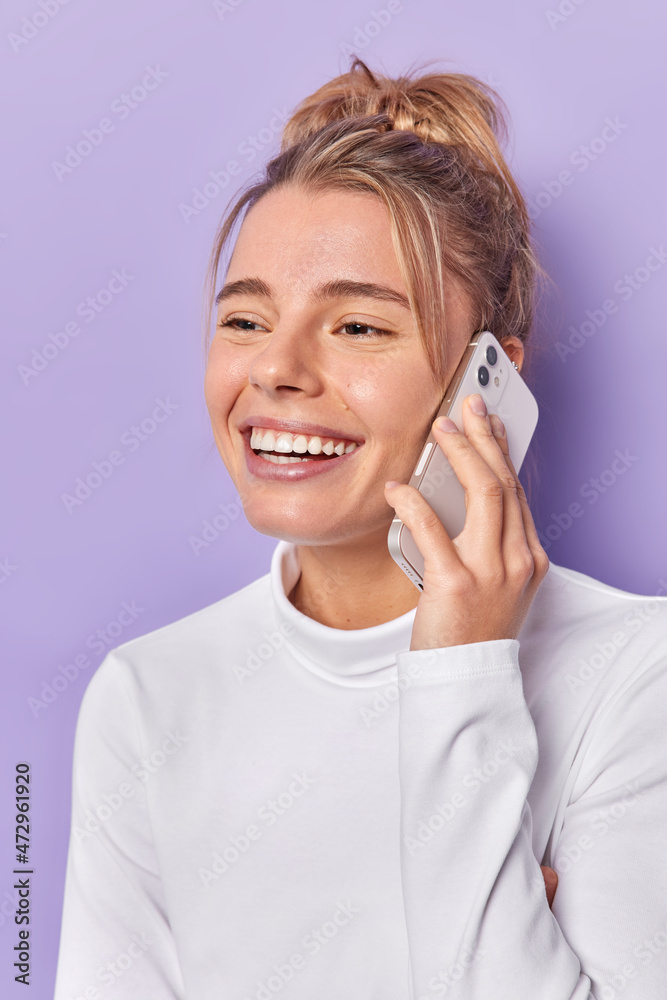 Vertical shot of happy young woman talks via mobile phone with best friend smiles gladfully enjoys nice conversation dressed in casual white jumper isolated over purple background. Communication Vertical shot of happy young woman talks via mobile phone with best friend smiles gladfully enjoys nice conversation dressed in casual white jumper isolated over purple background. Communication