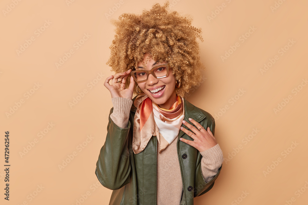 Happy woman in fashionable apparel keeps hand on spectacles smiles broadly glad to meet someone isolated over beige background. People lifestyle and emotions concept. Healthy cheerful person Happy woman in fashionable apparel keeps hand on spectacles smiles broadly glad to meet someone isolated over beige background. People lifestyle and emotions concept. Healthy cheerful person