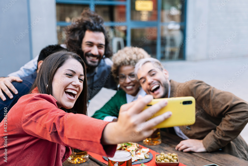 Obraz premium Young woman taking selfie photo of celebration with friends in outdoors bar to share on social media