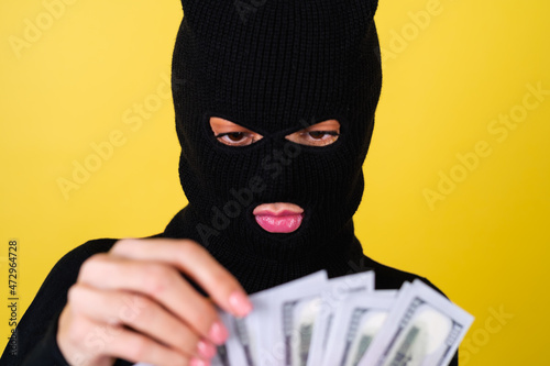 Young sexy beautiful woman with bright pink lips, balaclava on her head, holding a fan of 100 dollars, hacker, robbery, fraudster