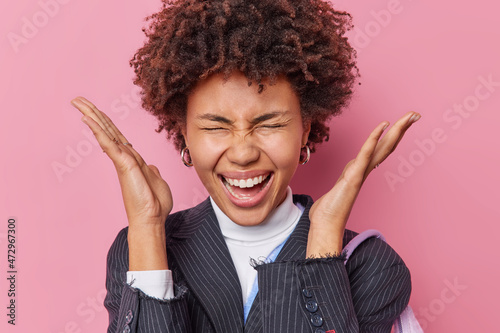 Positive emotional young pretty woman with curly hair raises palms near gead exclaims loudly keeps eyes closed wears black striped jacket reacts on wonderful news isolated over pink background. photo