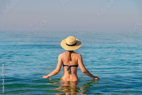 Back view of a slender caucasian young woman standing waist-deep in calm transparent sea water in a swimsuit and a straw hat
