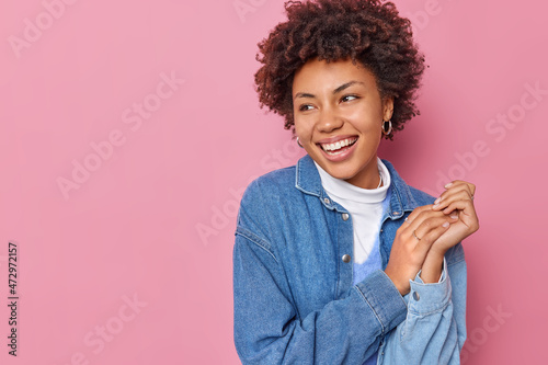 Studio shot of happy sincere young woman keeps hands together focused away with glad expression wears denim shirt looks joyfully at something funny isolated over pink background blank copy space