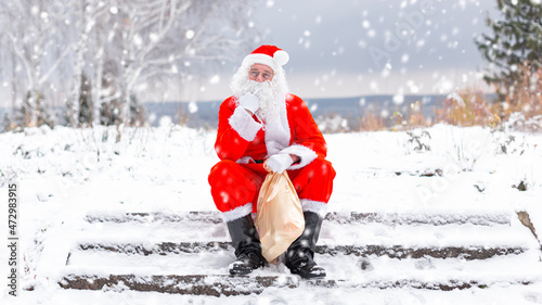 Tired and funny Santa Claus is sitting in nature in the forest, resting with a bag of gifts and wishes on Christmas and New Year's Eve